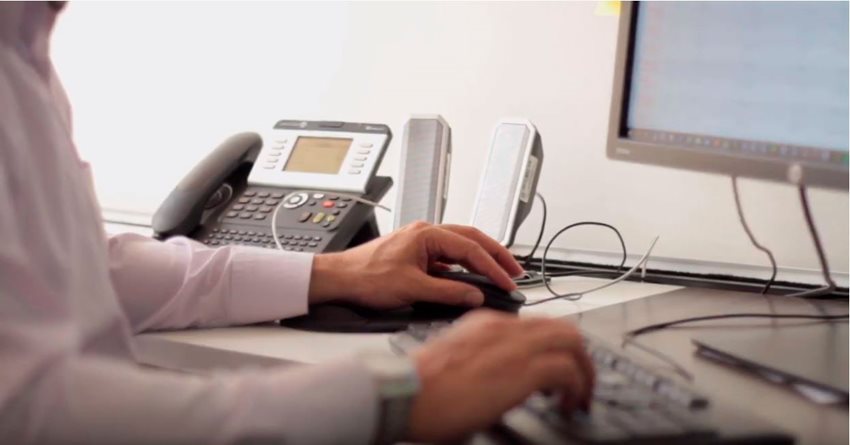 Manos de persona encima del teclado y del mouse de un computador, y al lado un teléfono.