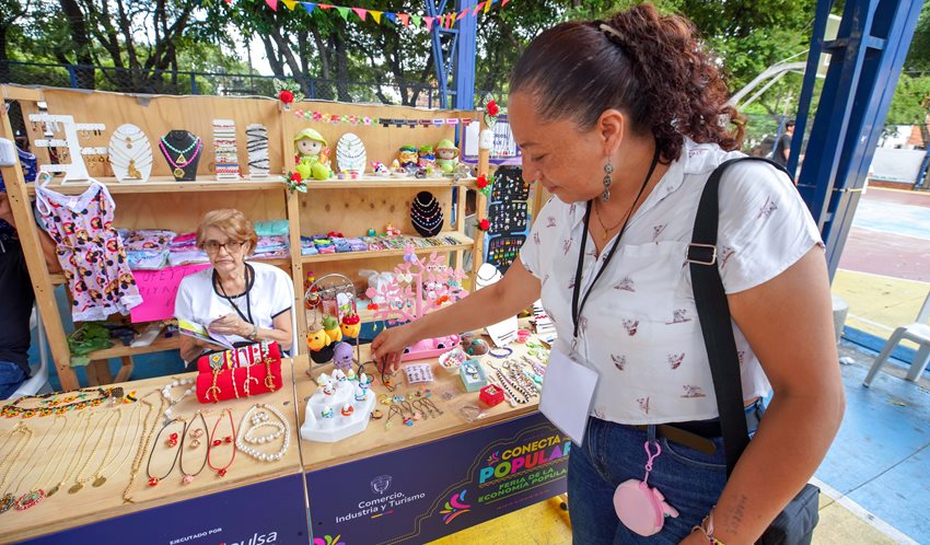 Descripción: Mujer mirando artesanías en estand de la Feria Conecta Popular, mientras la señora del lugar la observa.
