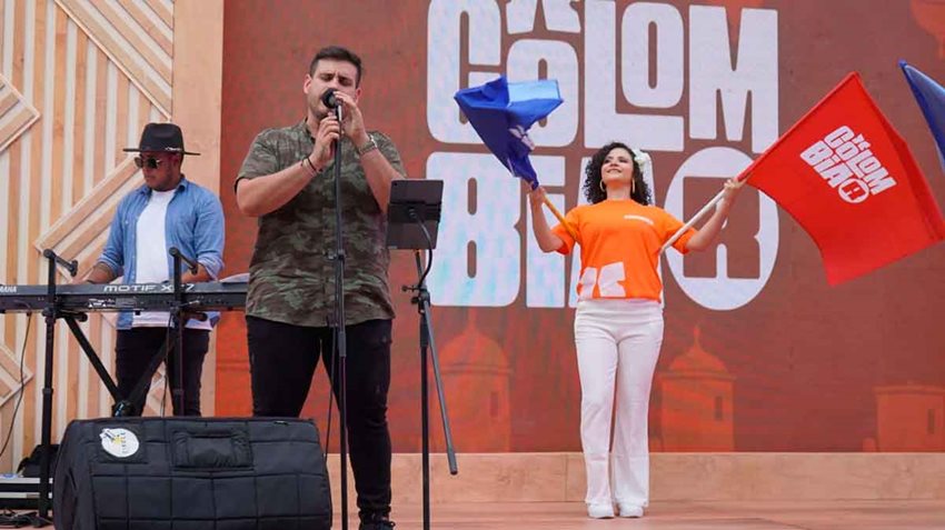 Hombre cantando en el escenario, con mujer en el fondo con banderas de colores y hombre tocando en el teclado.