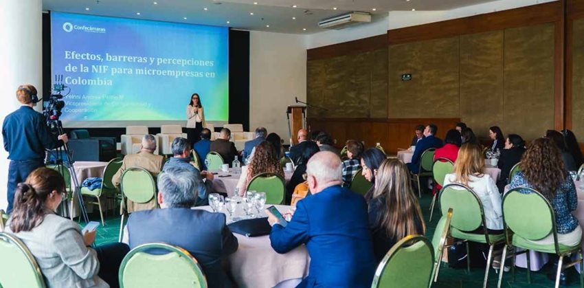 Descripción: Mujer hablando por micrófono y exponiendo al público asistente.