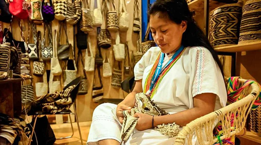 Mujer sentada, vestida de blanco, tejiendo mochila.