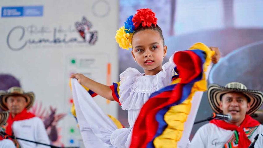 Niña morenita vestida con traje de cumbia blanco y decoraciones amarillo, azul y rojo, y detrás músicos.