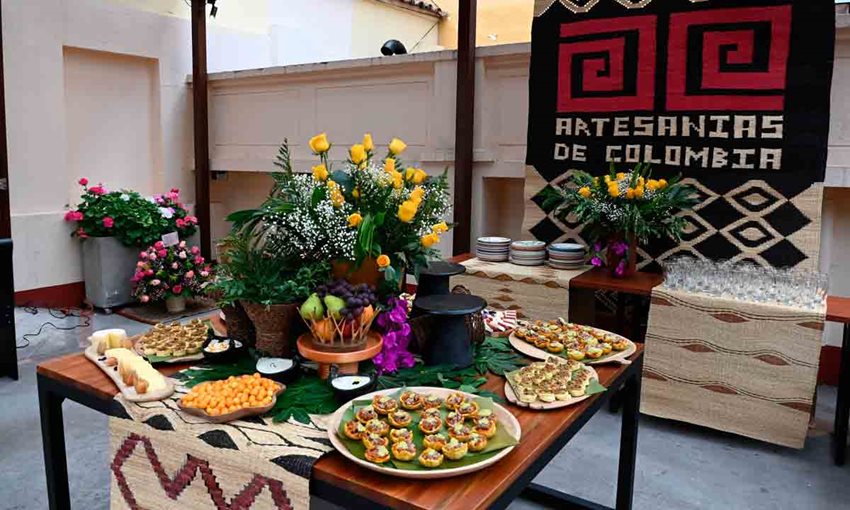 Foto de varios tipos de comida sobre platos, encima de una mesa, y al fondo el logo de Artesanía de Colombia.