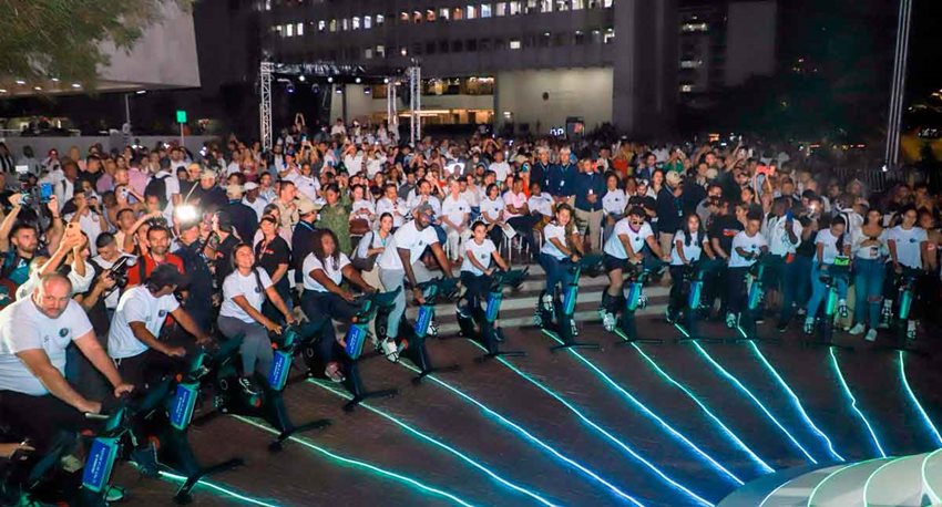 De noche, público mirando a personas con camisetas blancas, pedaleando en bicicletas estáticas que generan luz