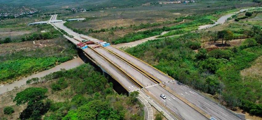 Foto panorámica de puente de tres carriles, con contenedores de colores atravesados, en medio de árboles.