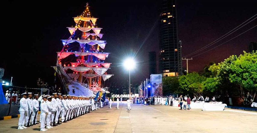 Hombres de la Armada, con uniforme blanco, formados al lado de un buque iluminado con colores de la bandera.
