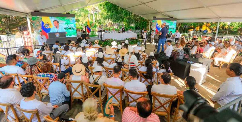 Foto panorámica de evento, con público sentado y con personas en tarima y el Presidente hablando en el atril.