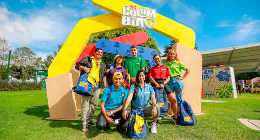 Descripción de la imagen: Cuatro mujeres y tres hombres, posando para la foto, con ropa de colores y frente al logo de Colombiar.