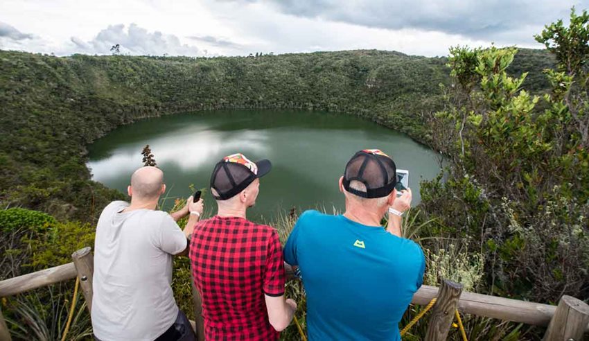 Tres hombres, dos con gorra, de espaldas, observando la laguna de Guatavita.