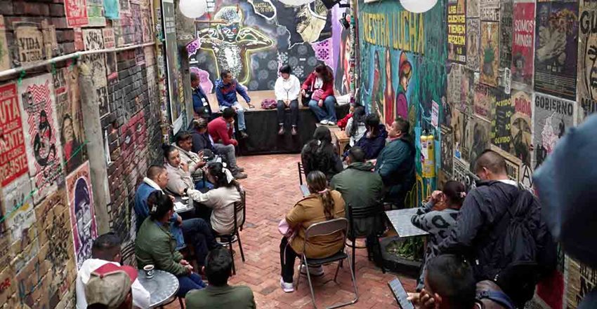 Descripción de la imagen: Hombres y mujeres compartiendo en mesas y de pie, en medio de una calle con grafitis.