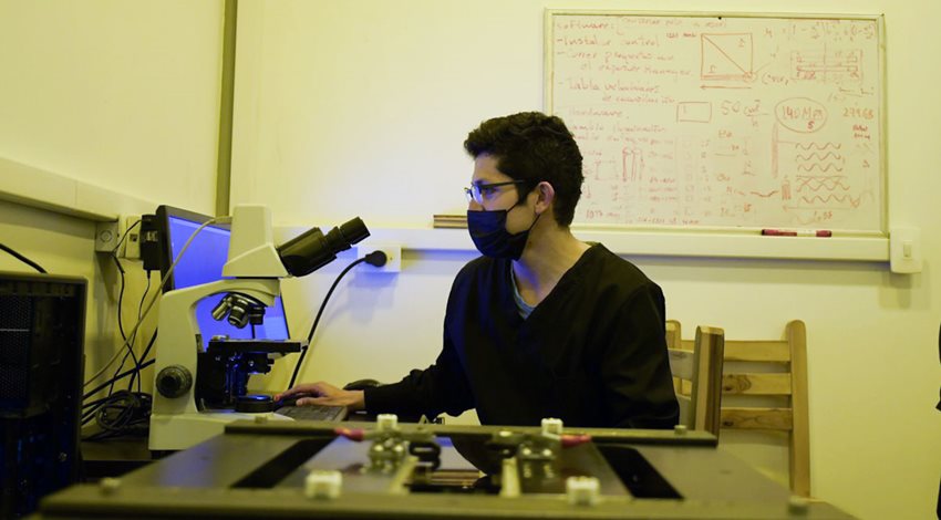 Joven con tapabocas frente a un microscopio y la pantalla de un computador.