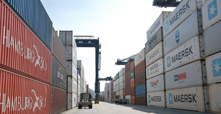 Contenedores de colores descargados en medio de una vía de zona franca y al lado una máquina de carga.