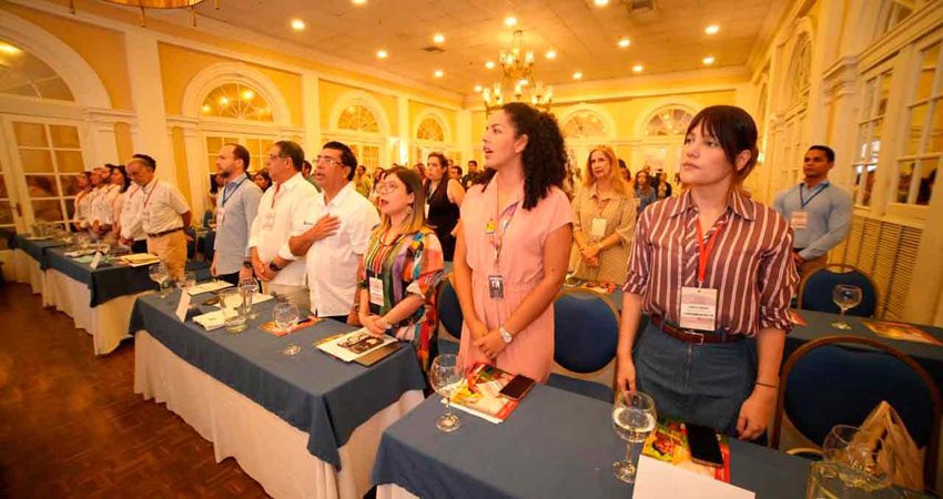 Hombres y mujeres de pie, dentro de un salón de conferencias, cantando el himno.