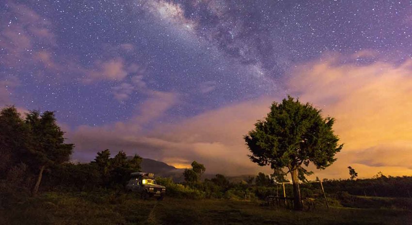 Descripción de la imagen: Paisaje del campo, con árboles y un carro en medio, junto con un cielo estrellado al atardecer.