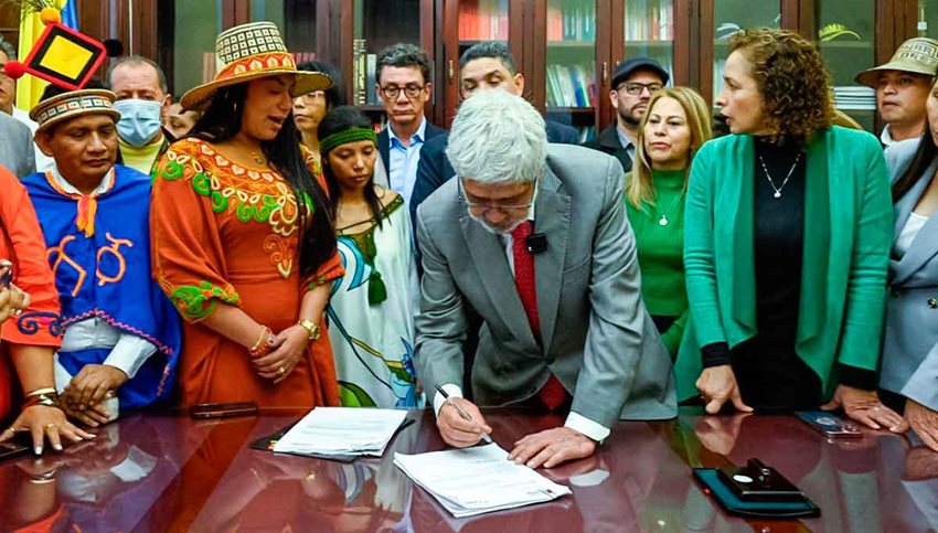 Ministro Germán Umaña rodeado de personas y firmando documentos encima de una mesa.