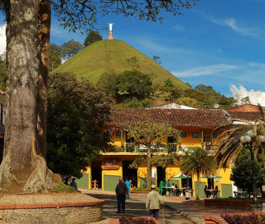Personas caminando en medio de un parque de un pueblo colombiano, con el fondo de una escultura de un Cristo.