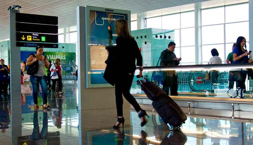 Descripción de la imagen: Mujer caminando con maleta de ruedas en medio de un aeropuerto, junto a personas alrededor que también caminan