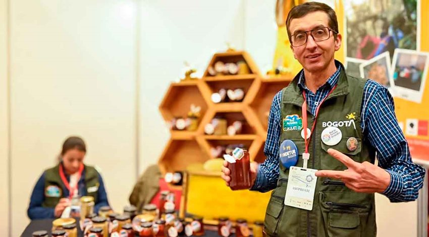 Hombre, con camisa azul y chaleco verde, posando para la foto con un tarro de vidrio en la mano.