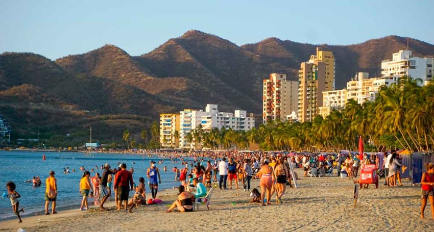Personas en la playa, al lado del mar, bañándose y tomando el sol, y de fondo edificios y montañas.
