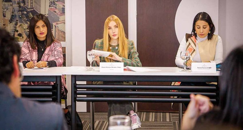 Ministra Diana Morales, sentada junto a dos mujeres, prestando atención a personas que hablan frente a ellas.