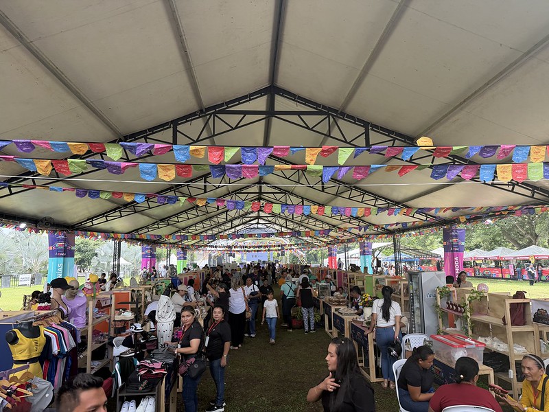 Descripción: Personas caminando por feria popular, con estands armados bajo una gran carpa blanca.