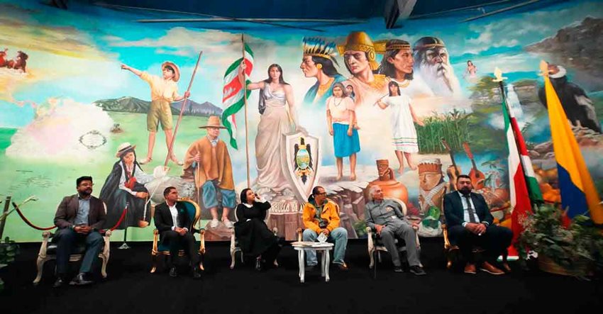 Una mujer y cinco hombres sentados exponiendo al público, con un mural representativo de Colombia al fondo.