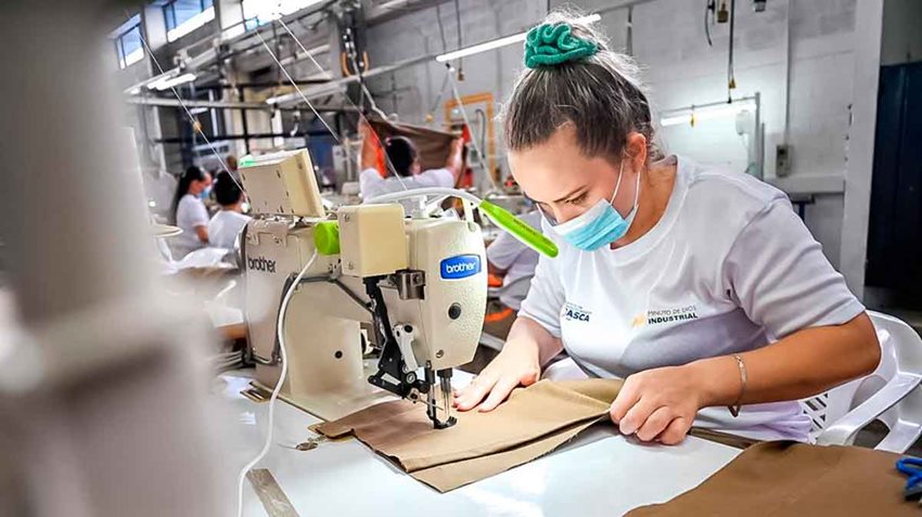 Mujer con tapabocas, camiseta blanca y cabello recogido, trabajando en un máquina de coser.