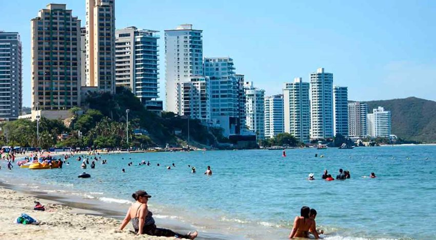 Descripción de la imagen: Personas tomando el sol en la playa, junto al mar, y de fondo edificios blancos.
