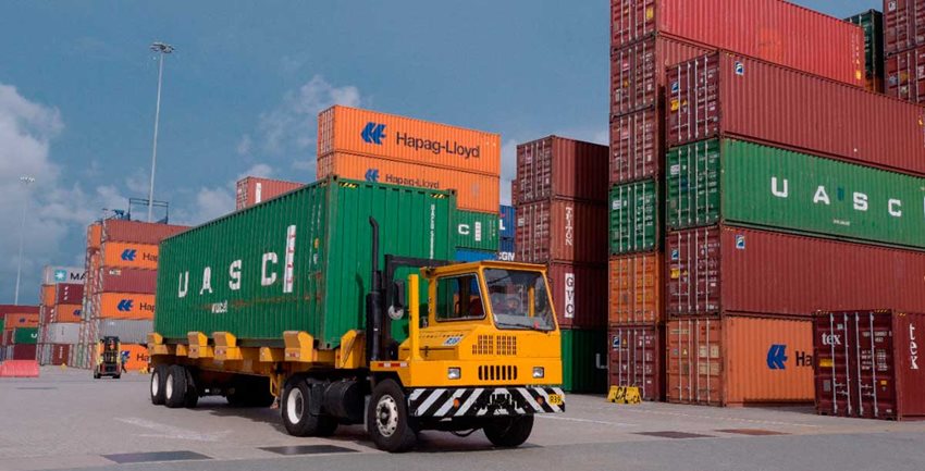 Camión verde con amarillo, transitando por en medio de contenedores de colores en puerto.