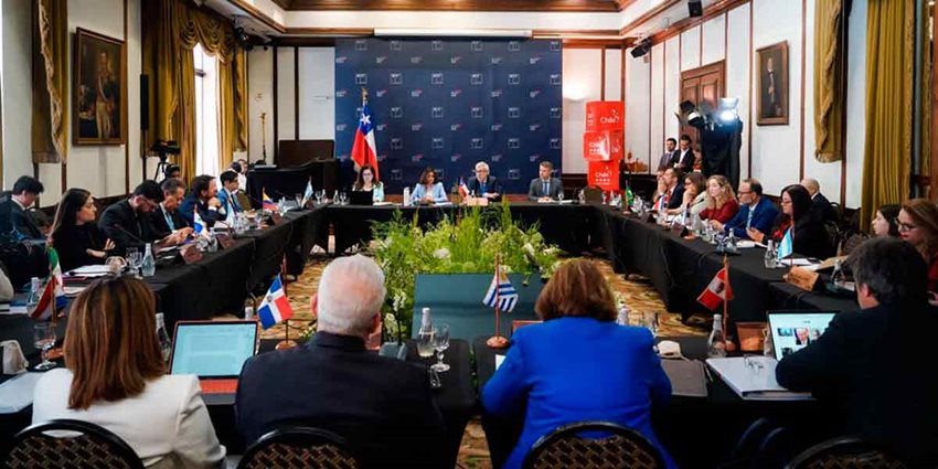 Hombres y mujeres, de traje, reunidos en mesa cuadrada dialogando.