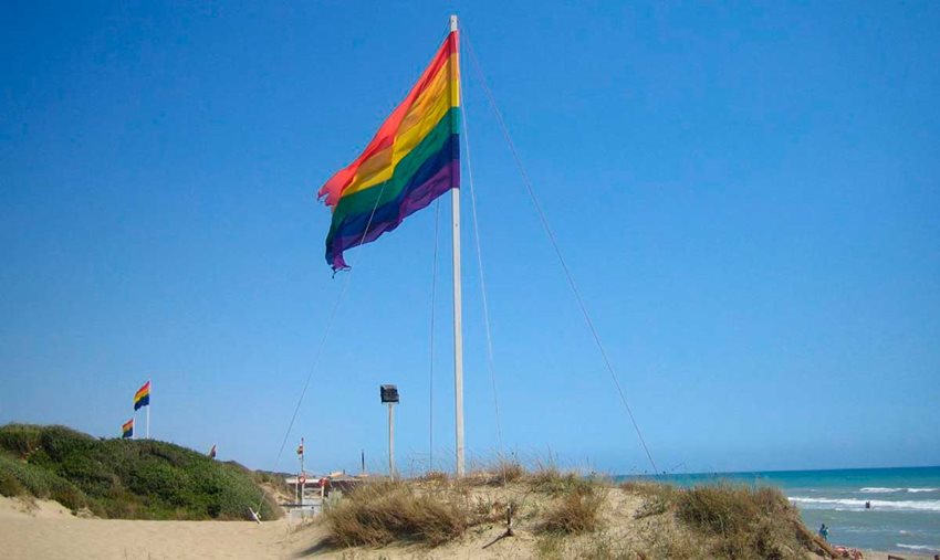 Descripción de la imagen: Bandera de colores arcoíris izada y ondeando en medio de la playa.