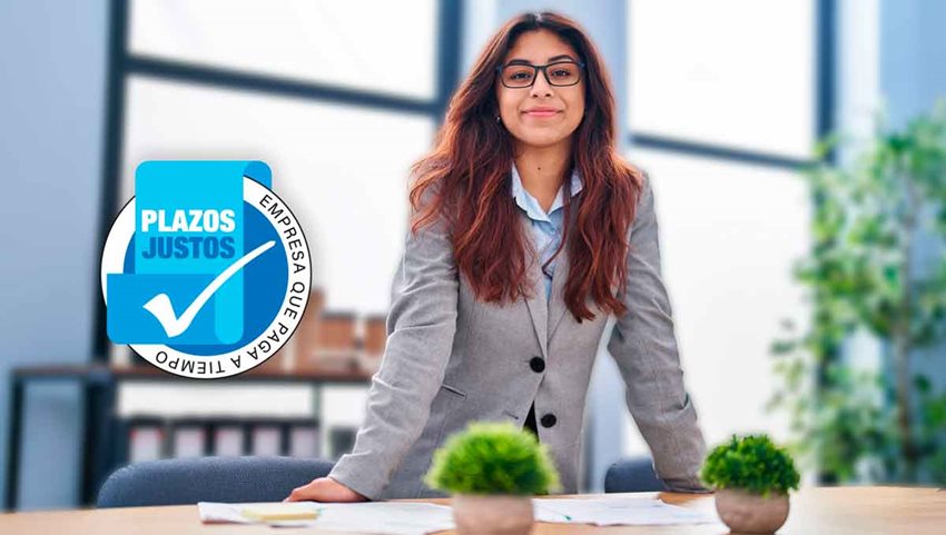 Mujer de cabello largo, con gafas y traje, posando para la foto, apoyando sus manos en un escritorio.