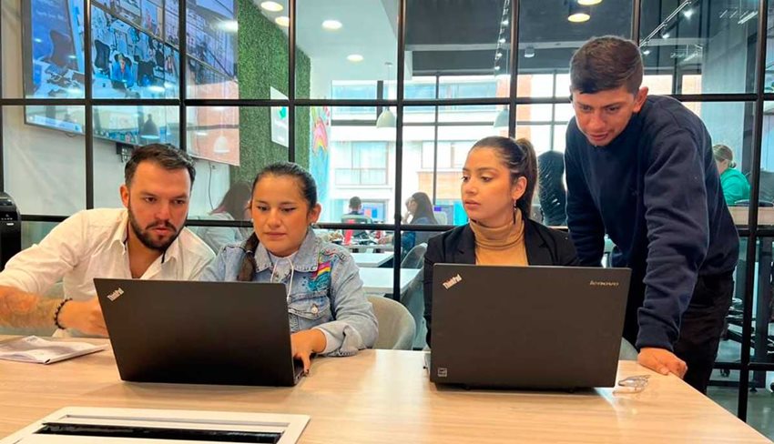 Dos hombres y dos mujeres, en una oficina, trabajando frente a dos computadores en medio de una reunión.