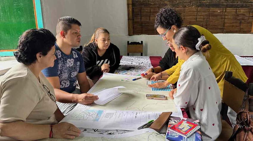 Personas reunidas alrededor de una mesa rectangular, mirando documentos en medio de una reunión.