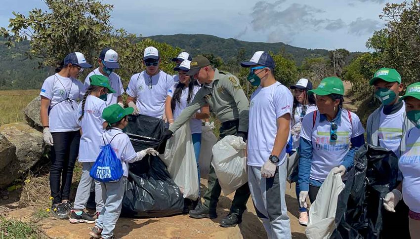 Personas con camisas blancas y gorras verdes de Colombia Limpia, junto a un policía, recogiendo basuras.