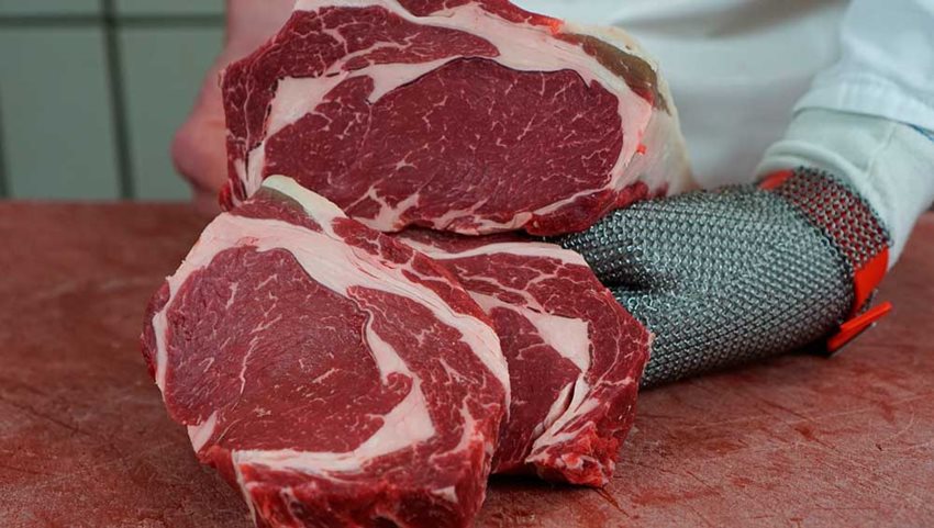 Pedazo de carne bovina sobre tabla, cortada a la mitad y sostenida por manos con guantes.