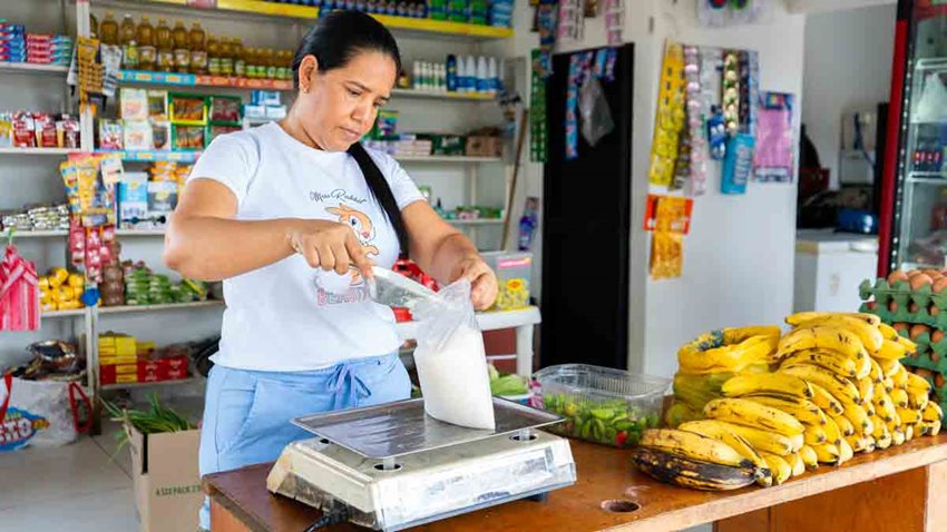 Mujer llenando y pesando una bolsa de azúcar en medio de una tienda de barrio.