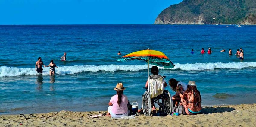 Mujer en silla de ruedas, en la arena, junto a otras mujeres sentadas en la arena, observando el mar.