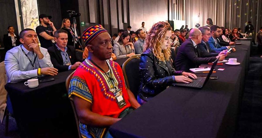 Descripción de la imagen: Hombres y mujeres sentados en sillas y recostados con sus brazos en mesas, prestando atención al frente.