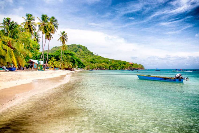 Paisaje de playa, con palmeras y una lancha en el mar, y al fondo motañas.