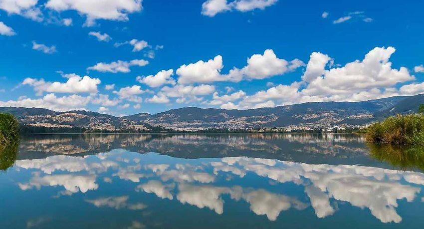 Descripción de la imagen: Paisaje, de día, de un lago con montañas en el fondo y el reflejo de éstas en el lago.