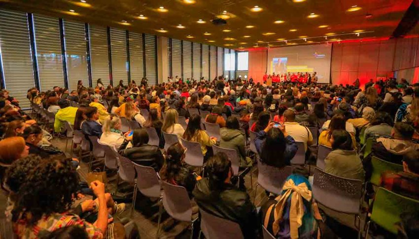 Muchas personas sentadas en sillas en un auditorio, prestando atención a quienes están en la tarima hablando.