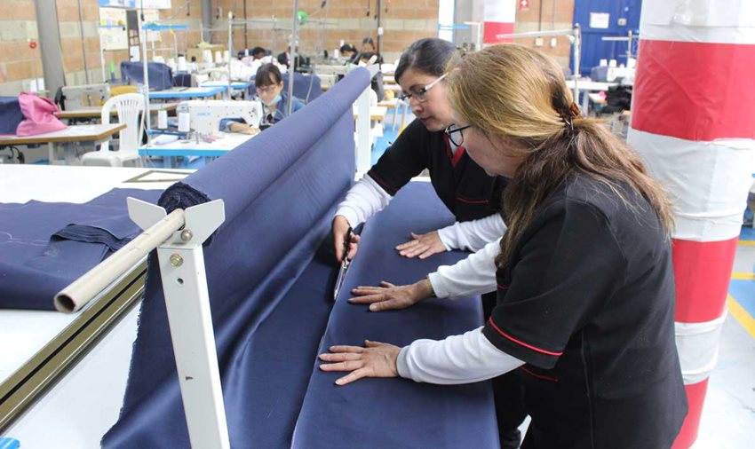 Dos mujeres, con uniforme negro, cortando tela azul oscura con unas tijeras sobre una máquina.