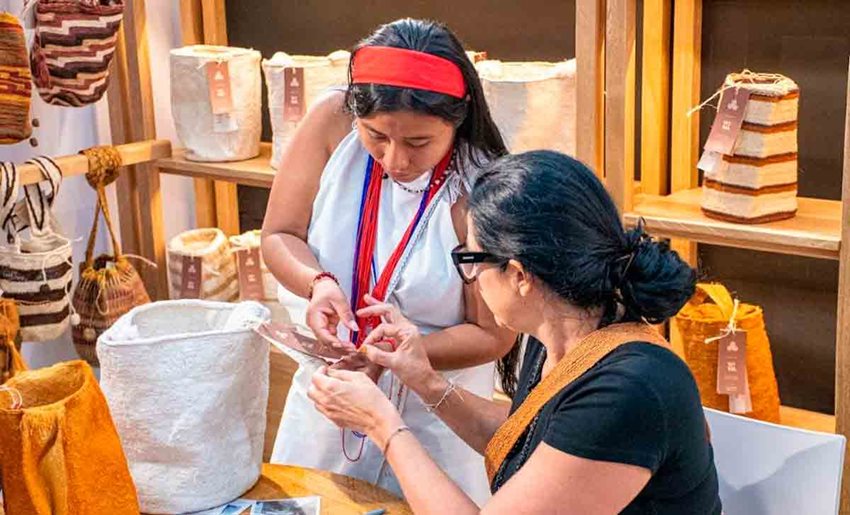 Mujer indígena mostrando artesanías a otra mujer con gafas y vestida de negro.