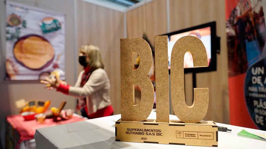 Galardón en madera del logo BIC y al fondo una señora exponiendo su emprendimiento en su estand.