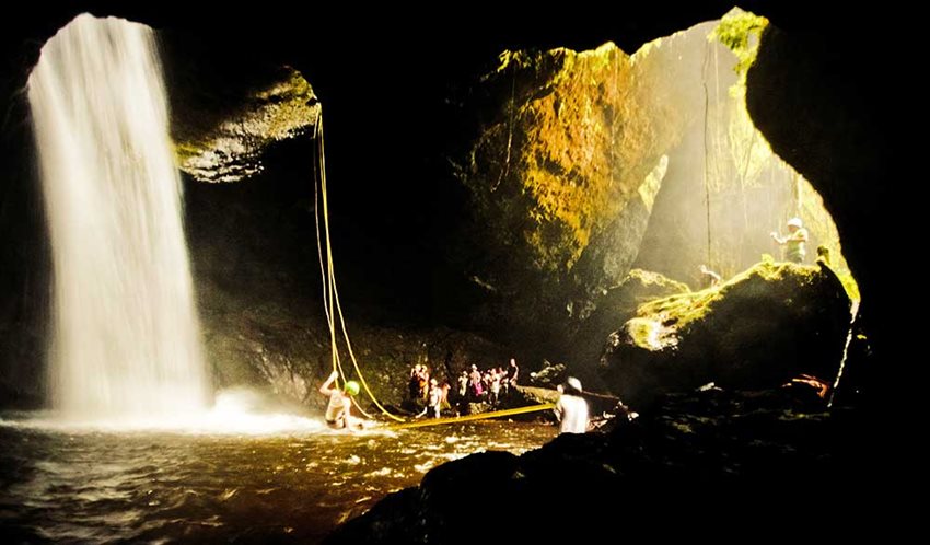 Guías y turistas dentro de una cueva, con cascada, explorándola.