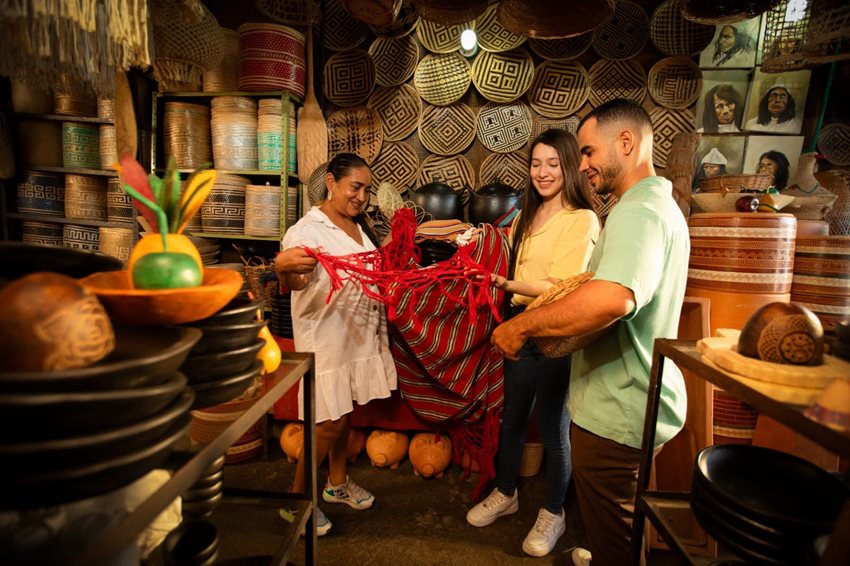 Descripción: Dos mujeres y un hombre mirando una hamaca roja, en medio de un cuarto lleno de artesanías.