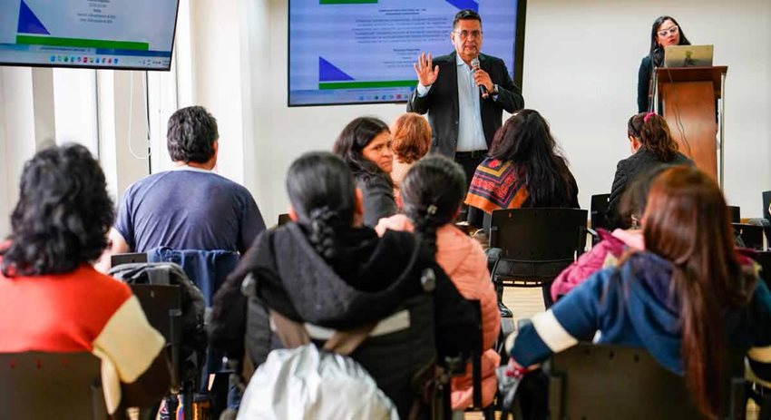 Hombre hablando por micrófono al público asistente, personas que están sentadas en sillas.