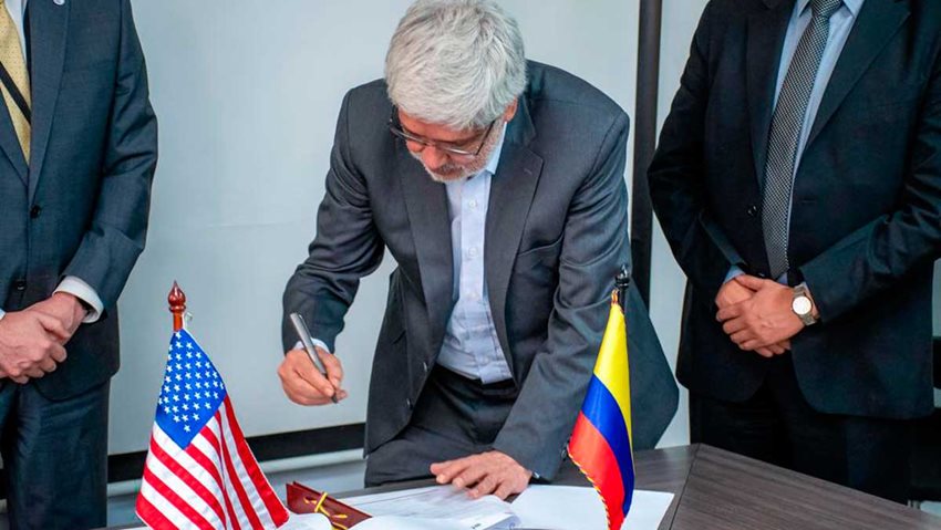 Descripción de la imagen: Ministro Germán Umaña firmando documentos puestos sobre una mesa, al lado de las banderas de USA y Colombia.