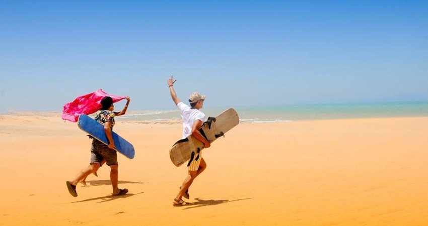 Descripción de la imagen: Tres personas corriendo en la arena, con patinetas en la mano, hacia el mar.
